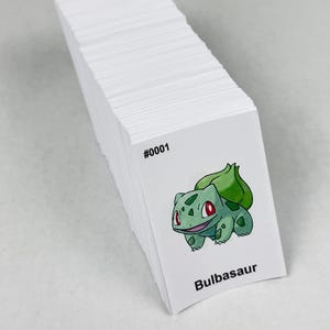 May include: A stack of rectangular white cards featuring a printed illustration of a green Bulbasaur character with a bulb on its back. The text #0001 is printed in black above the character, and the name Bulbasaur is printed in black below it.