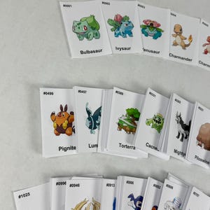 May include: A collection of white rectangular trading cards featuring colorful illustrations of various creatures. Each card displays a unique identification number, a character name, and a specific creature graphic, arranged in rows on a plain surface.