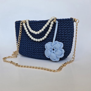 May include: Navy blue crochet handbag with a gold chain strap and pearl accents. A light blue crochet flower is attached to the front. The bag has a rectangular shape.