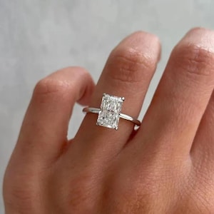 May include: A radiant-cut diamond ring with a thin, silver band. The rectangular diamond is the focal point, reflecting light. The ring is shown on a finger, highlighting its elegant design and sparkle.