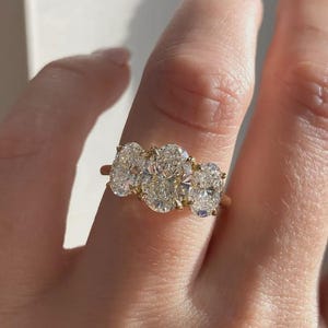 May include: A gold ring with three oval-cut diamonds. The diamonds are clear and set in a row on a thin gold band. The ring is displayed on a finger, showcasing the jewellery's design and sparkle.