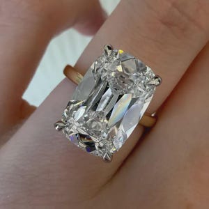 Peut inclure: Une bague ornée d'une grande pierre transparente de taille coussin rectangulaire aux coins arrondis, fixée par quatre griffes argentées. L'anneau de la bague est en métal doré lisse et poli, contrastant avec la monture argentée.