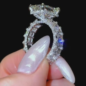 May include: A silver-toned ring featuring a large, rectangular-cut center stone set in a prong setting. The band is encrusted with multiple smaller, oval-shaped stones that extend around the entire circumference, creating a highly reflective, sparkling appearance against a dark background.