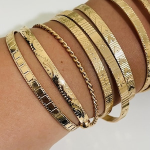 Op de afbeelding: Een set van zeven goudkleurige metalen armbanden die samen worden gedragen. De armbanden hebben verschillende texturen en patronen, waaronder een gedraaid touwontwerp, geometrische vierkanten, gegraveerde bloemmotieven en herhalende driehoeksvormen.
