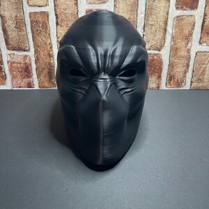 May include: A black, full-face mask with a textured surface. The mask has eye and nose openings and is set against a brick wall background.