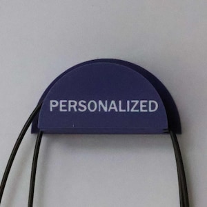 Pode incluir: Uma etiqueta azul escuro, semicircular, com a palavra "PERSONALIZED" em letras brancas. Dois cordões pretos estendem-se da parte inferior da etiqueta, contra um fundo branco.