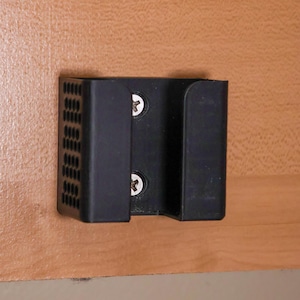 May include: A black plastic wall-mounted holder, secured with two screws, is attached to a light brown wooden surface. The holder has a rectangular shape with a series of small, evenly spaced holes on the left side.
