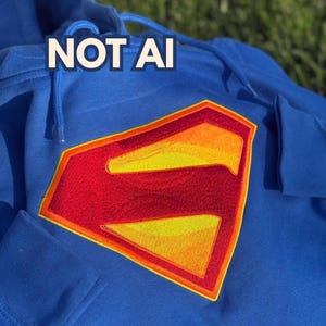 May include: A bright blue hooded sweatshirt featuring a large, embroidered diamond-shaped emblem in red and yellow on the chest. The emblem displays a stylized letter S. The text NOT AI is printed in white with a dark outline above the emblem.