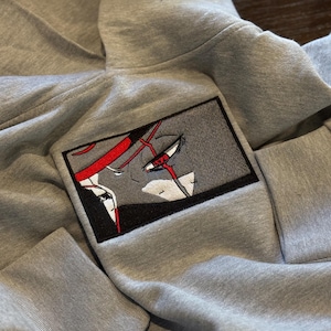 Peut inclure: Un sweat-shirt à capuche gris clair avec un écusson rectangulaire brodé sur la poitrine. L'écusson présente un gros plan stylisé de style anime d'un visage avec des accents rouges, des contours noirs et des reflets blancs, bordé de noir.