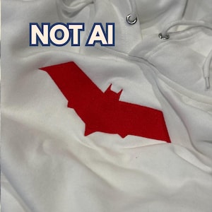 Könnte beinhalten: Ein weißer Kapuzenpullover mit einem großen, gestickten roten Fledermaus-Emblem auf der Brust. Der Text NOT AI ist in Blau und Weiß über dem Emblem aufgedruckt. Der Hoodie verfügt über eine Kapuze mit Kordelzug sowie silbernen Metallösen.