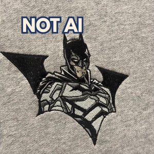 May include: A heather grey fabric featuring an embroidered Batman character design in black, white, and grey thread. Above the character, the text NOT AI is embroidered in bold blue letters. The design is centred on the textured grey textile surface.