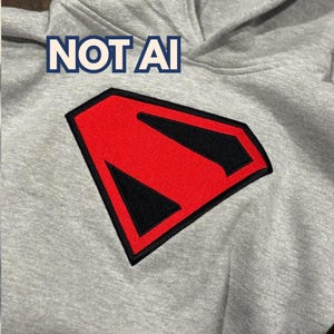以下が含まれることがあります： 胸元に大きな赤と黒のダイヤモンド型刺繍エンブレムがあしらわれたライトグレーのフード付きスウェットシャツ。エンブレムの上には、濃い青色の太字で「NOT AI」というテキストが生地にプリントされています。