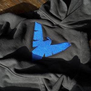 Könnte beinhalten: Ein anthrazitfarbener Kapuzenpullover mit einem auffälligen, leuchtend blauen, gestickten vogelartigen Emblem in der Mitte der Brust. Der Stoff hat eine weiche Textur und das Design zeigt eine stilisierte Vogelsilhouette mit kantigen Flügeln.