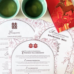 May include: A traditional Chinese tea ceremony setup. Two teal tea cups filled with green tea are placed near red envelopes with horse illustrations and printed ceremony guides. The guides feature text and floral designs.