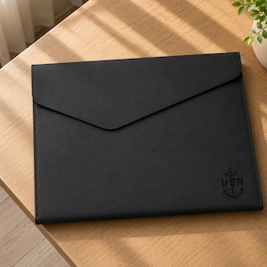 May include: A black rectangular document folder with an envelope-style flap closure. The front surface features a debossed emblem of an anchor with the letters USN positioned directly above it in the lower right corner of the folder.