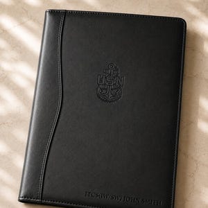 May include: A black rectangular portfolio with a smooth finish and stitched edges. The front features an embossed USN anchor emblem in the center and the text ITCS(IW/SW) JOHN SMITH debossed in the bottom right corner.