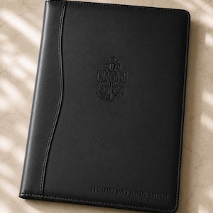 May include: A black rectangular portfolio with a smooth finish and stitched edges. The cover features an embossed USN anchor emblem in the center and the text ITC(IW/SW) JOHN SMITH debossed in the bottom right corner.