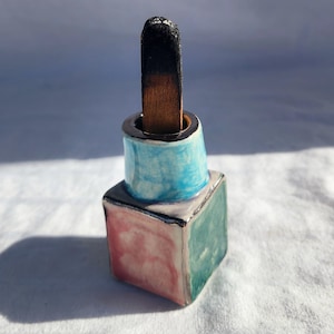 Può includere: Un piccolo porta incenso in ceramica con una glassa blu, verde e rosa. Un bastoncino di incenso in legno sta bruciando all'interno del porta incenso.