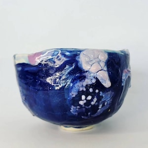 Pode incluir: Uma tigela de cerâmica artesanal com uma superfície texturizada. A tigela apresenta um esmalte azul escuro com detalhes brancos, rosa e azul claro. A borda da tigela tem uma forma irregular e orgânica, e a base é de cor creme claro.