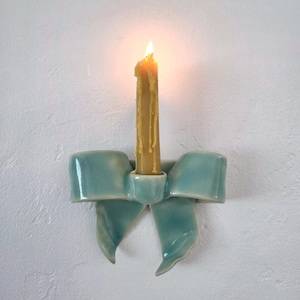 May include: A light blue ceramic wall sconce shaped like a decorative bow with a central holder containing a single lit yellow taper candle. The candle features visible wax drips running down its sides, and the ceramic bow has a glossy finish.