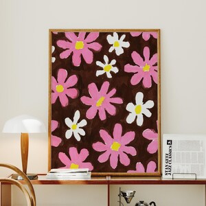 May include: A framed floral art print featuring a repeating pattern of pink and white flowers with yellow centers on a dark brown background. The artwork is displayed on a wooden shelf with other decorative items.