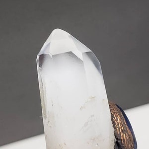 May include: A clear quartz crystal point with a faceted tip. The crystal is mounted on a base with a dark blue and bronze-colored finish. The background is a neutral gray.