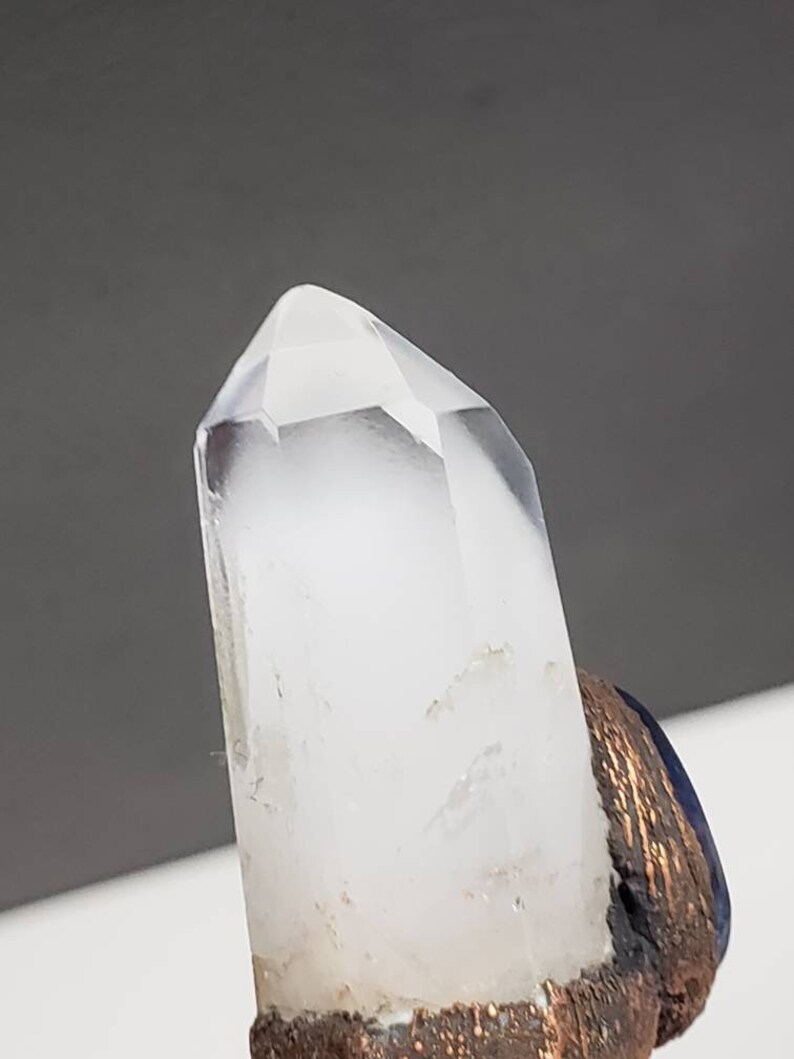 May include: A clear quartz crystal point with a faceted tip. The crystal is mounted on a base with a dark blue and bronze-colored finish. The background is a neutral gray.
