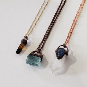 May include: Three necklaces with different gemstone pendants. The first has a brown crystal, the second a blue-green crystal, and the third a clear crystal with a blue stone. Each pendant is attached to a chain.