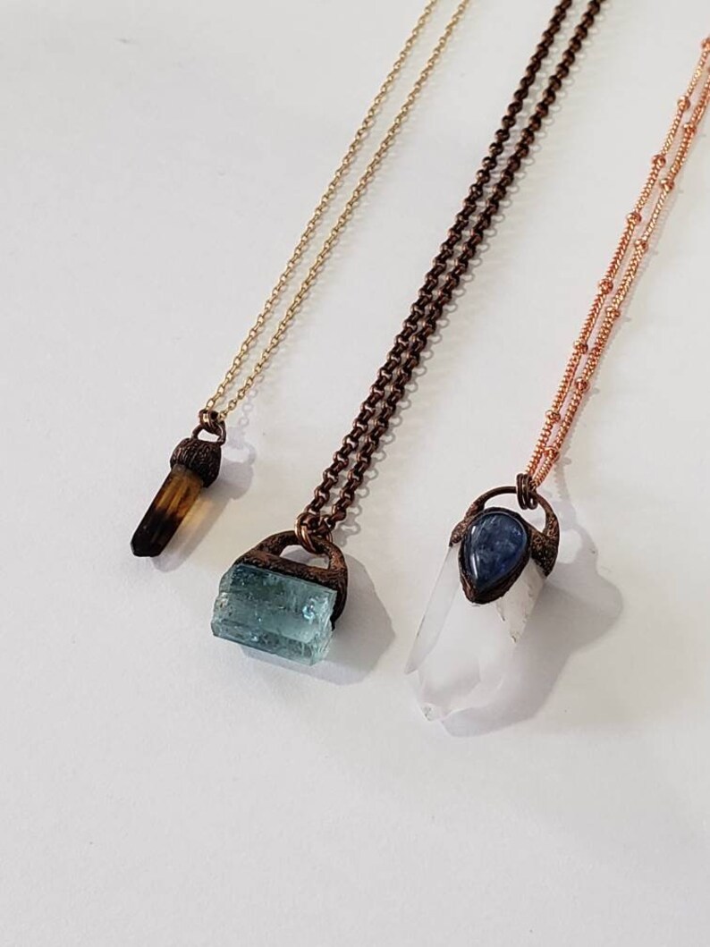 May include: Three necklaces with different gemstone pendants. The first has a brown crystal, the second a blue-green crystal, and the third a clear crystal with a blue stone. Each pendant is attached to a chain.