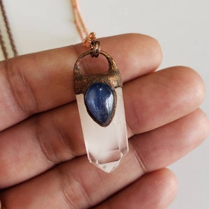 May include: A clear quartz crystal pendant with a blue gemstone accent, suspended from a copper-colored chain. The crystal is faceted and has a pointed tip. The pendant is held in place by a copper-colored metal setting.