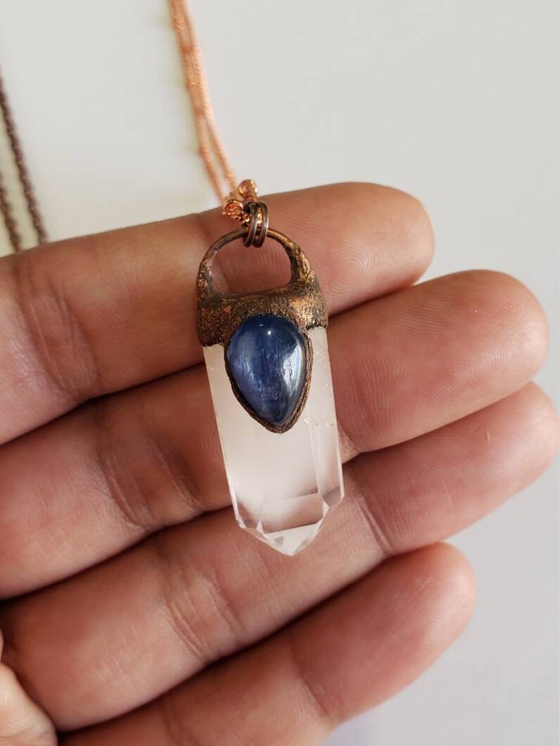 May include: A clear quartz crystal pendant with a blue gemstone accent, suspended from a copper-colored chain. The crystal is faceted and has a pointed tip. The pendant is held in place by a copper-colored metal setting.