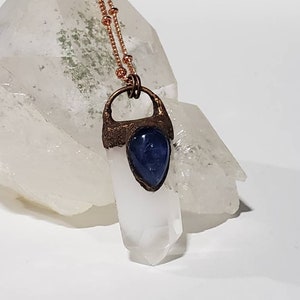 May include: A copper-colored necklace with a clear quartz crystal pendant and a blue gemstone accent. The pendant is set in a textured copper frame and hangs from a delicate beaded chain. The necklace is displayed on a white surface.