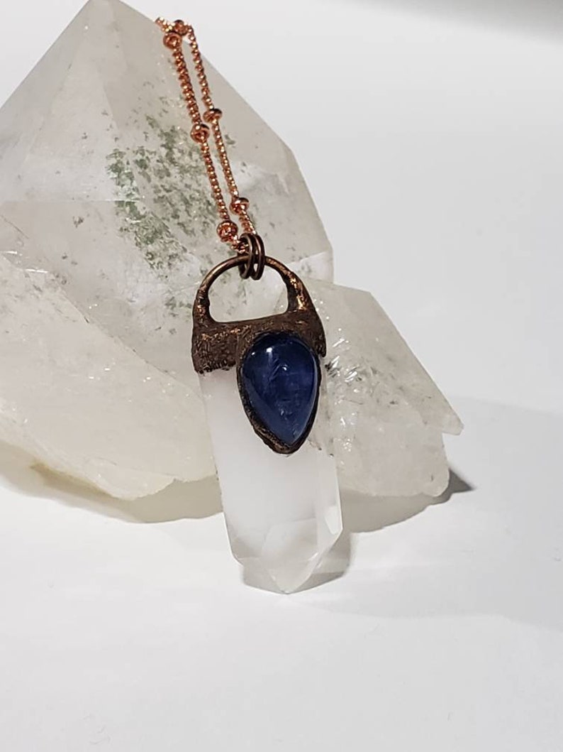May include: A copper-colored necklace with a clear quartz crystal pendant and a blue gemstone accent. The pendant is set in a textured copper frame and hangs from a delicate beaded chain. The necklace is displayed on a white surface.