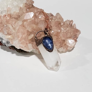 May include: A clear quartz crystal pendant necklace with a copper-colored chain. The crystal is capped with a blue stone and copper-colored metal. The background features a cluster of pinkish-white crystals.