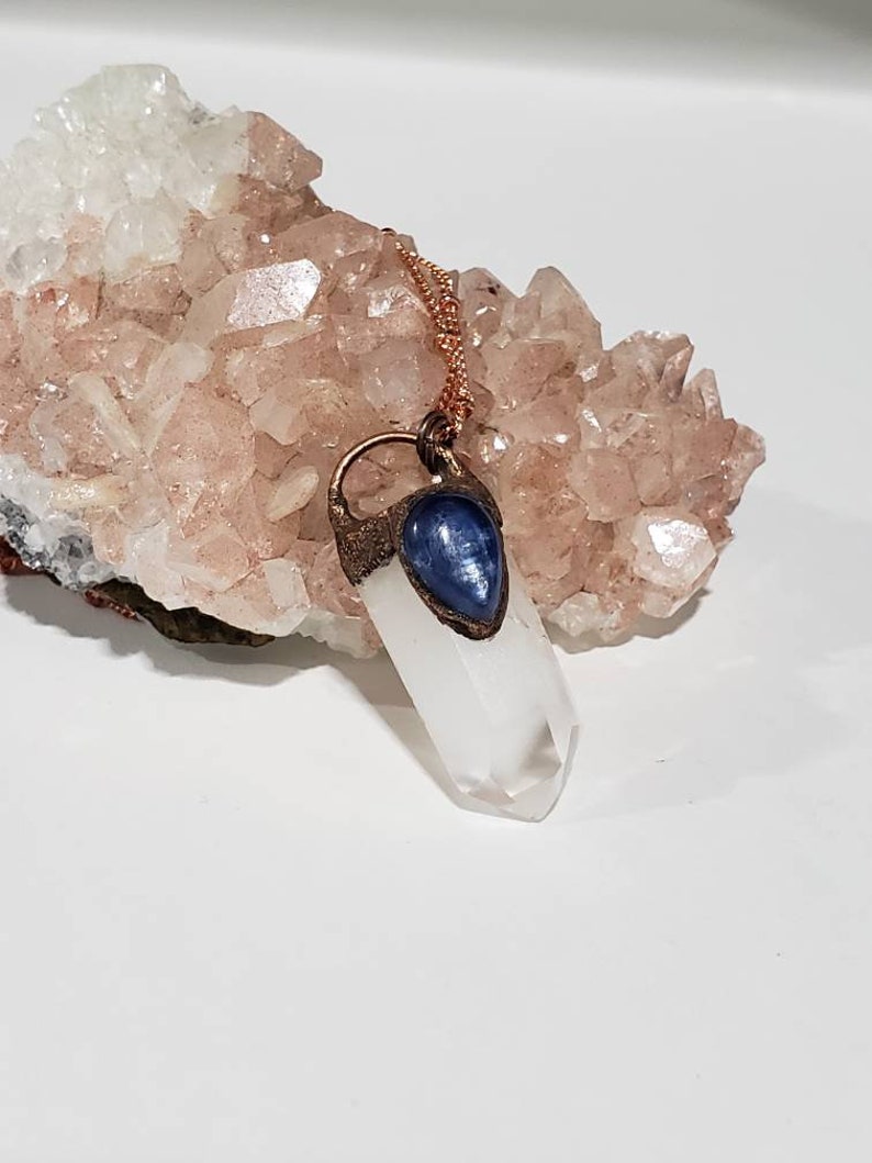 May include: A clear quartz crystal pendant necklace with a copper-colored chain. The crystal is capped with a blue stone and copper-colored metal. The background features a cluster of pinkish-white crystals.