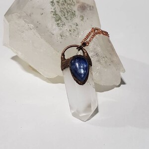 May include: A clear quartz crystal pendant with a blue gemstone accent, suspended from a copper chain. The pendant is set against a larger, rough-cut quartz crystal, creating a contrast in textures and shapes. The overall aesthetic is natural and earthy.