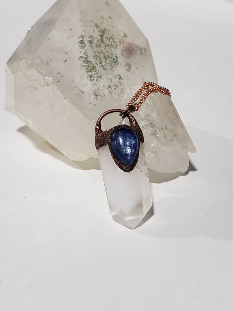 May include: A clear quartz crystal pendant with a blue gemstone accent, suspended from a copper chain. The pendant is set against a larger, rough-cut quartz crystal, creating a contrast in textures and shapes. The overall aesthetic is natural and earthy.