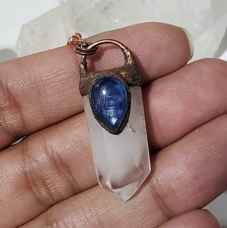 May include: A clear quartz crystal pendant with a blue kyanite stone accent. The pendant is set in a copper-colored metal and hangs from a delicate copper chain. The crystal is approximately 2 inches long.