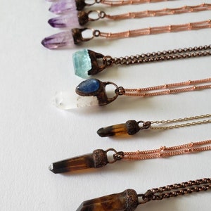 May include: An assortment of crystal pendant necklaces. The necklaces feature various crystals, including amethyst, aquamarine, and smoky quartz, set in copper-colored metal. The chains are also copper-colored. The crystals are in different shapes and sizes.