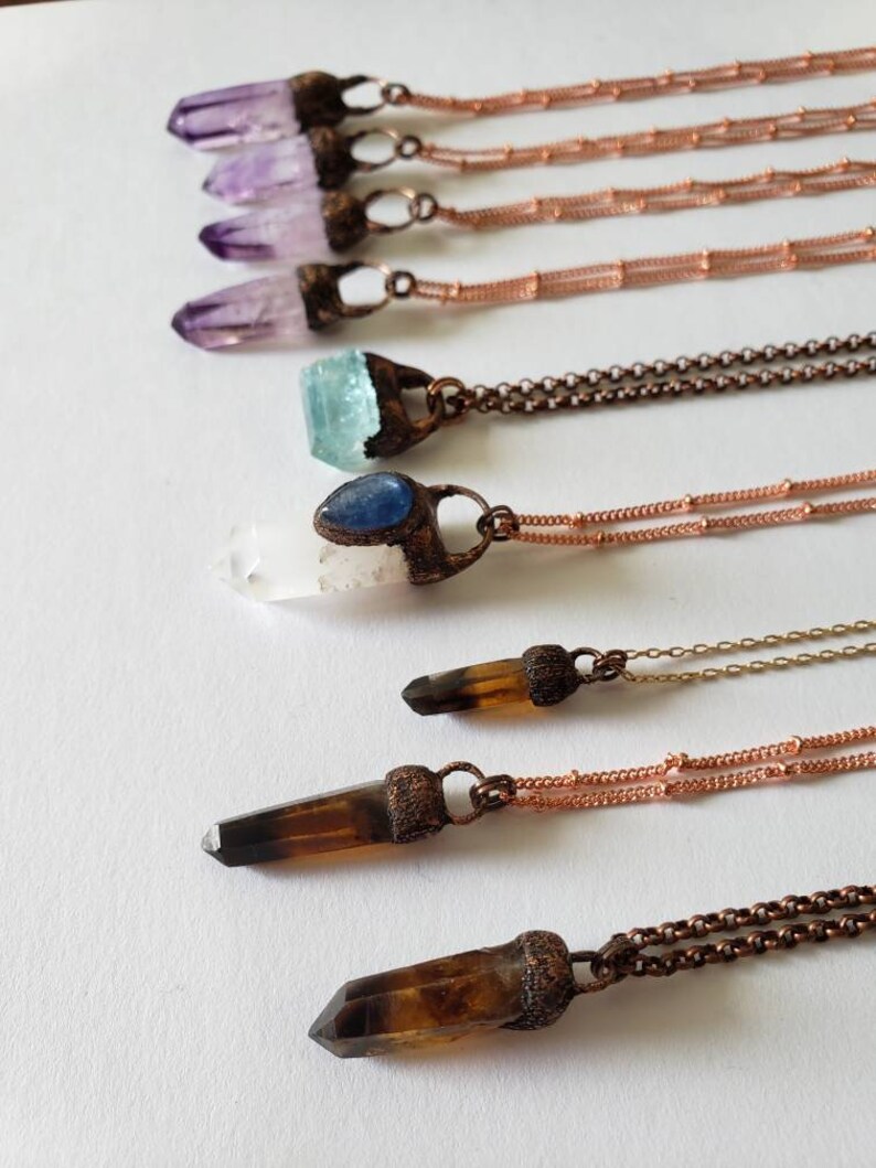 May include: An assortment of crystal pendant necklaces. The necklaces feature various crystals, including amethyst, aquamarine, and smoky quartz, set in copper-colored metal. The chains are also copper-colored. The crystals are in different shapes and sizes.