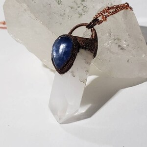 May include: A copper-colored pendant necklace featuring a clear quartz crystal point. The pendant includes a teardrop-shaped blue stone. The necklace chain is also copper-colored. The crystal is set against a white background.