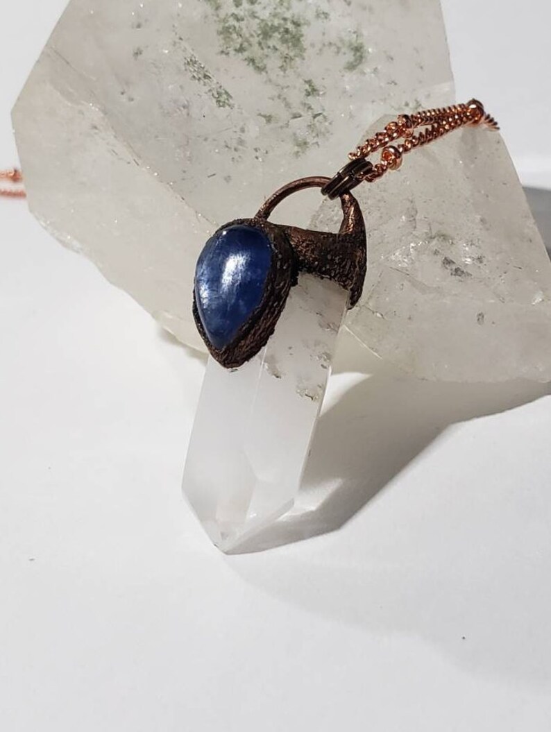 May include: A copper-colored pendant necklace featuring a clear quartz crystal point. The pendant includes a teardrop-shaped blue stone. The necklace chain is also copper-colored. The crystal is set against a white background.