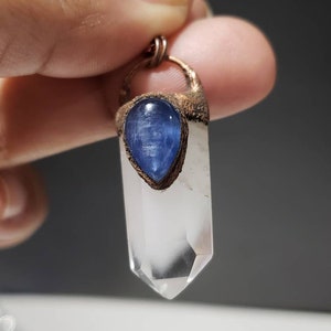 May include: A clear quartz crystal pendant with a blue kyanite stone and copper-colored metal accents. The crystal is a long, pointed shape. The blue stone is teardrop-shaped. The pendant is suspended from a thin, brown cord.
