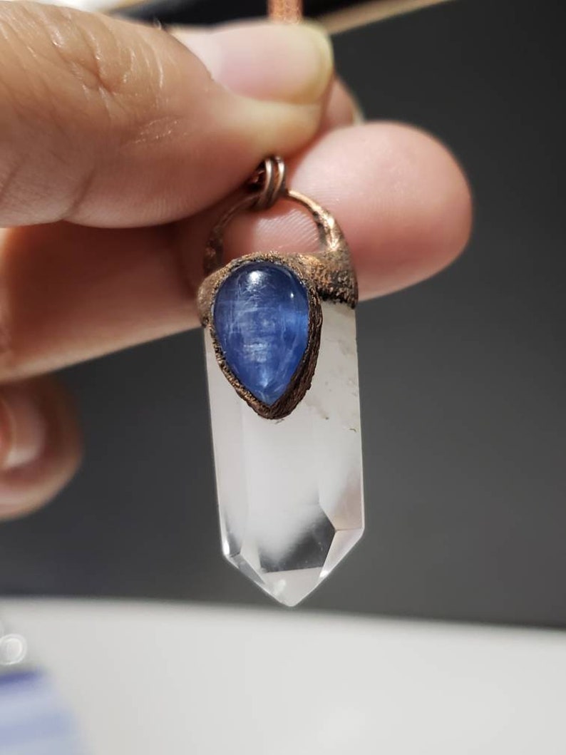 May include: A clear quartz crystal pendant with a blue kyanite stone and copper-colored metal accents. The crystal is a long, pointed shape. The blue stone is teardrop-shaped. The pendant is suspended from a thin, brown cord.