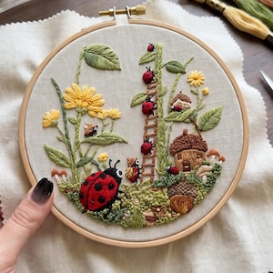 Puede incluir: Un bastidor de bordado circular de madera que contiene un panel de tela con un diseño detallado de naturaleza. El bordado presenta una mariquita roja grande, una escalera con mariquitas pequeñas, flores amarillas, follaje verde y una casa de bellota.
