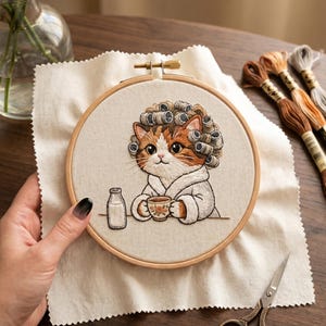 May include: An embroidery hoop features a cat wearing curlers and a robe, holding a teacup. A small milk bottle sits beside the cat. The embroidery is on a cream-colored fabric, with brown and orange thread. Scissors and embroidery floss are also visible.