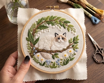 May include: An embroidered hoop art piece featuring a fluffy cat resting on a branch, surrounded by green leaves, white flowers, and blue accents. The cat has a white and brown coat. Embroidery floss and scissors are also visible.