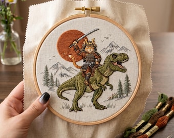 May include: Embroidered hoop art depicting a cat samurai on a green T-Rex, with a sunset, mountains, and pine trees in the background. The cat wears armour and holds a sword. The hoop is held in a hand.