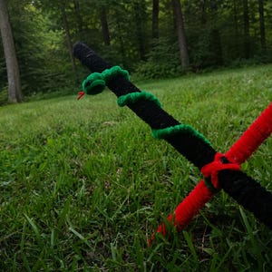 May include: A craft project made of fuzzy pipe cleaners featuring a long black and red rod crossed with another red rod. A small green snake with a red tongue is coiled around the upper section of the black rod, featuring two small black eyes.