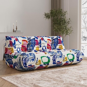 May include: A low-profile sofa with a vibrant, abstract pattern. The upholstery features a white background with bold strokes of blue, pink, red, yellow, and green. The sofa has a rounded shape and is placed in a well-lit room.